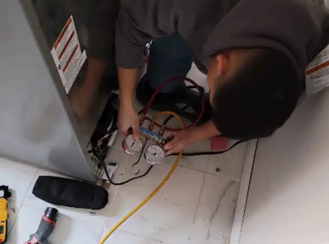 An Elite Appliance technician kneeling behind a pulled-out refrigerator with refrigerant gauges connected to the sealed system, tool bag and drill on the floor
